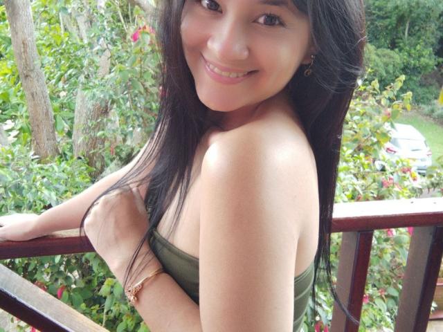 SwetShastity with long black hair, smiling, green strapless top, showing her shoulder and arm, wearing a gold bracelet, standing on a wooden balcony, greenery in background.