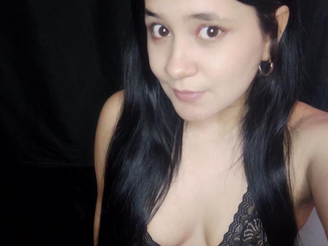 SwetShastity with long black hair, wearing a lace top that reveals her small tits, and gold hoop earrings, has a subtle smile and brown eyes. She's looking at the camera.