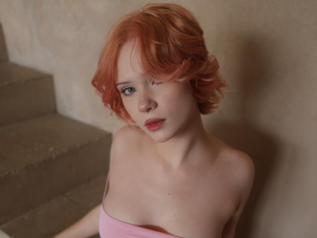 MollyWettmond with red curly hair, pale skin, and pink strapless top, showing her small tits and red lips, sitting on concrete stairs, looking up with a slight smile.
