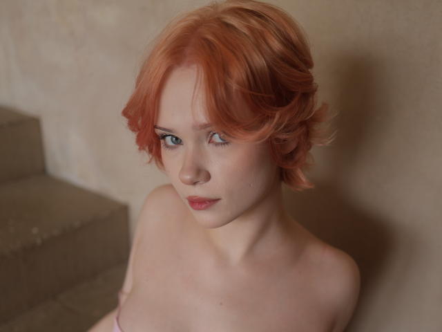 MollyWettmond has short, wavy, orange-red hair, pale skin, and blue eyes, with a serious expression, topless, showing small, bare breasts, sitting on concrete stairs.