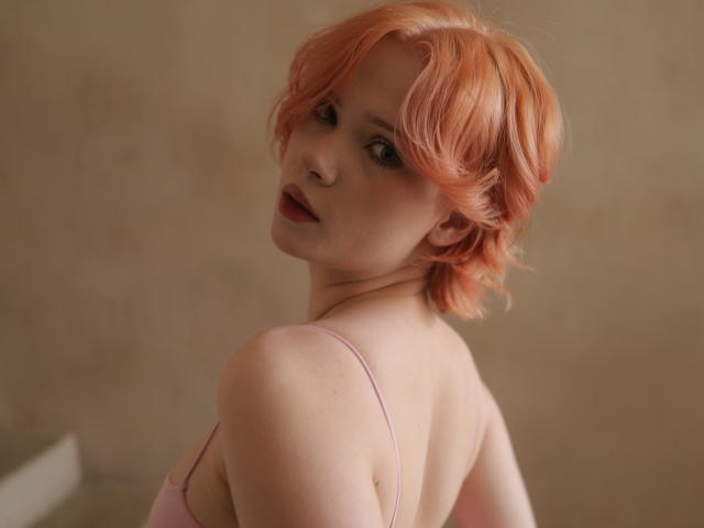MollyWettmond with pink, wavy hair looking over her shoulder, wearing a pink, thin-strapped top, showing her bare, soft shoulders and slightly visible, small tits.