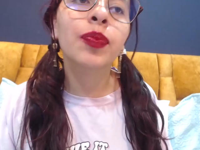 Julia Waine mit vollen, roten Lippen, Brille und Zöpfen, trägt ein weißes T-Shirt und zeigt ihren leicht geöffneten Mund und ihre kleine Nase. Im Hintergrund ist ein gelber Kopfteil zu sehen..