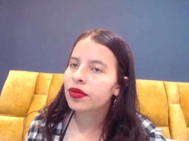 JuliaWaine has red lipstick, black hair, and is wearing a checkered shirt, sitting on a yellow, tufted, leather-like seat with a blue wall behind her.