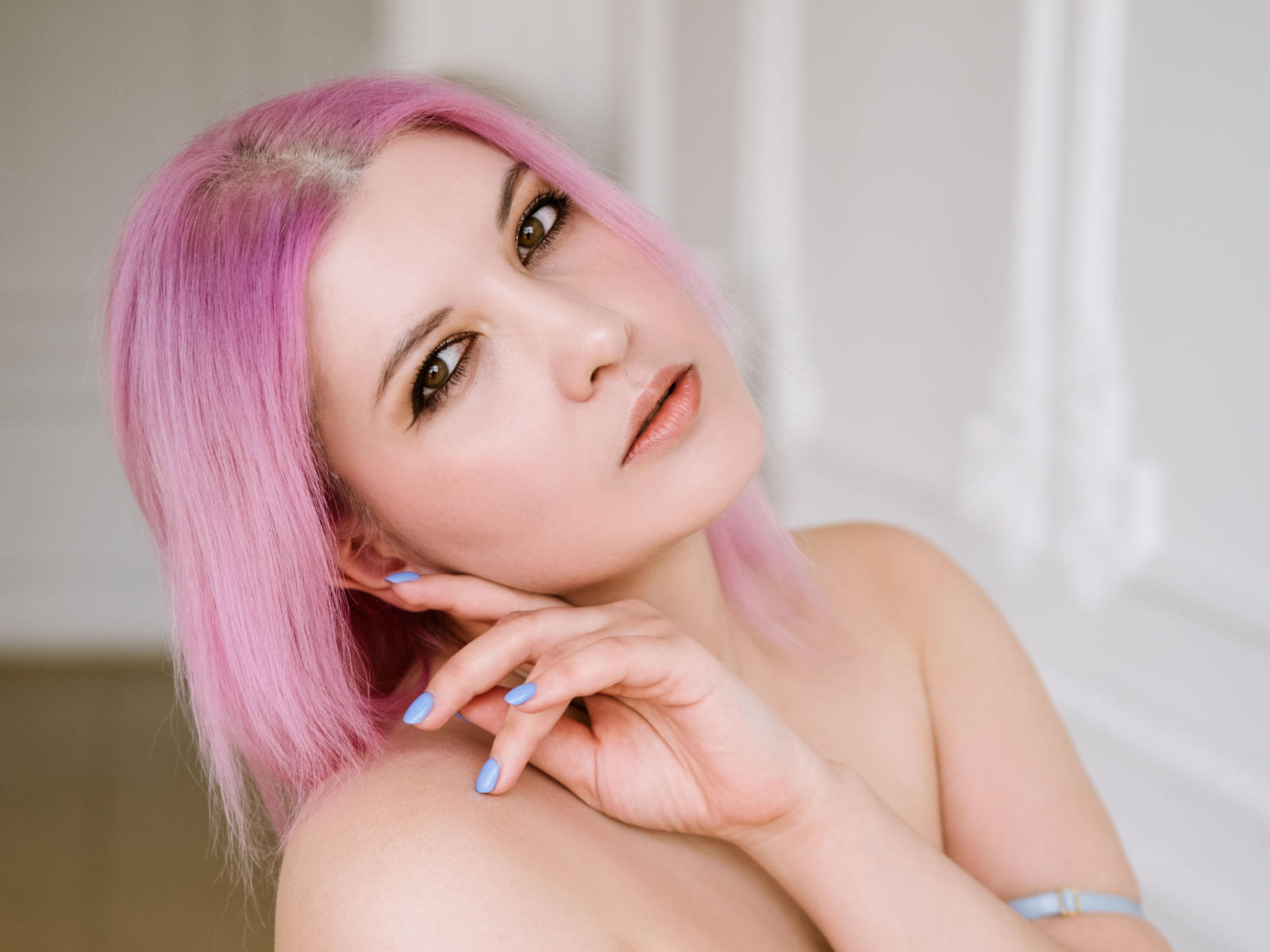 OliSunnyS tiene el pelo rosa, piel clara, ojos marrones y está sin ropa, con uñas azules, tocándose suavemente el cuello, mirando a la cámara con una sonrisa sutil..