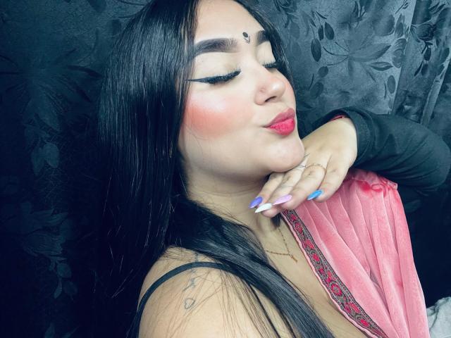AnaShalimarr, con lunghi capelli neri, rossetto rosa e unghie dipinte di blu-rosa, bacia il suo braccio con le maniche nere, indossa una maglietta rosa e ha un bindi sulla fronte..