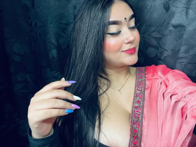 AnaShalimarr mit langen, schwarzen Haaren, einem pinken Oberteil, lila-blauen-weißen Nägeln, rotem Lippenstift, dunklen Augenbrauen und einem kleinen Punkt auf der Stirn, lächelt leicht und hält ihre Haare..