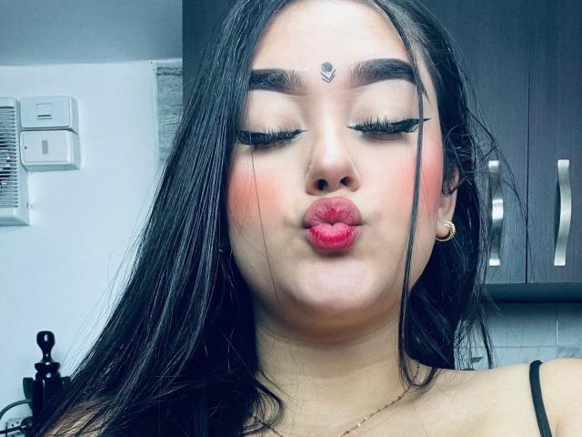 AnaShalimarr mit langem, schwarzem Haar, rotem Lippenstift und dicker, schwarzer Eyeliner, die ihre vollen, rosafarbenen Lippen zu einem Schmollmund formt, trägt ein kleines Herz auf ihrer Stirn, in einer Küche..