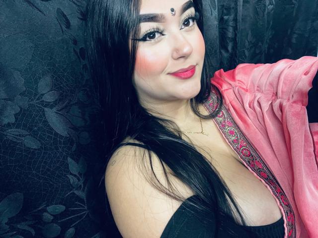 AnaShalimarr, con cabello negro largo, lápiz labial rojo y un chal rosa, mostrando sus senos con una blusa negra, con un bindi en la frente, sonriendo a la cámara..