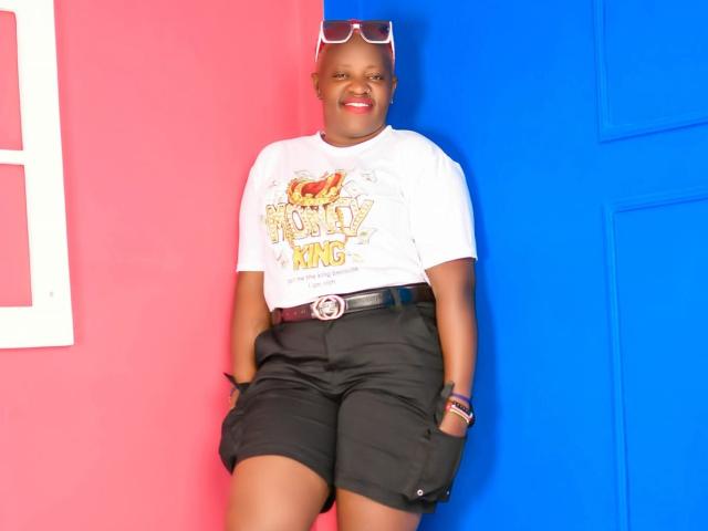 Ebonyalya mit dunkler Haut, rotem Lippenstift, weißem T-Shirt, schwarzen Shorts, Gürtel mit runder Schnalle, Sonnenbrille auf dem Kopf, lächelnd, stehend vor rosa und blauen Wänden..