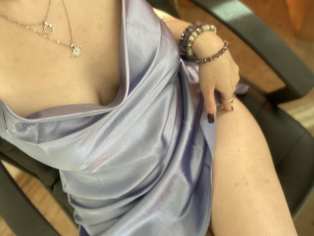 Khori's tits are visible in a silky, purple dress with a deep cleavage. She's wearing a butterfly necklace and bracelets, sitting on a black chair with her leg exposed.