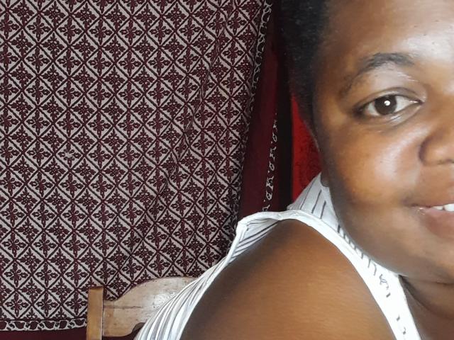 DoraKatty's brown face, small smile, dark eyes, and black curly hair in a white striped top, showing her shoulder and part of her chest. Background: red and white patterned fabric.