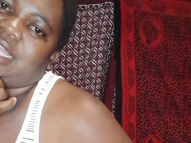 DoraKatty with dark skin, short curly hair, brown eyes, smiling, white tank top, hand on neck, lying on patterned pillow, red and black background, looking at camera.
