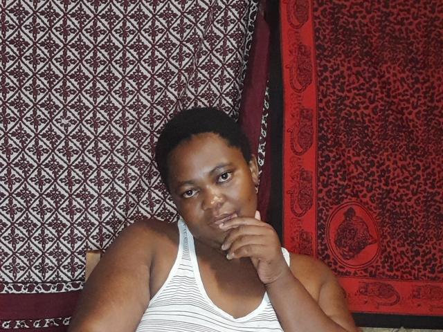 DoraKatty with dark skin, short curly hair, wearing a white striped tank top, resting her chin on her hand, looking at the camera with a slight smile.