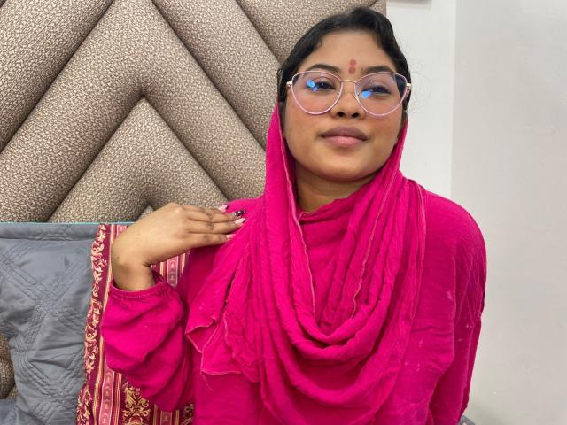 ShailaYojana with dark skin, pink glasses, and pink headscarf, wearing a pink dress, has a forehead dot and a calm expression, holding her scarf with pink nails.