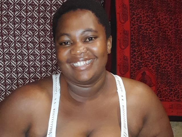 DoraKatty with dark skin, short curly hair, smiling, wearing a white tank top, showing her big ass and small tits, sitting in front of a red patterned background.