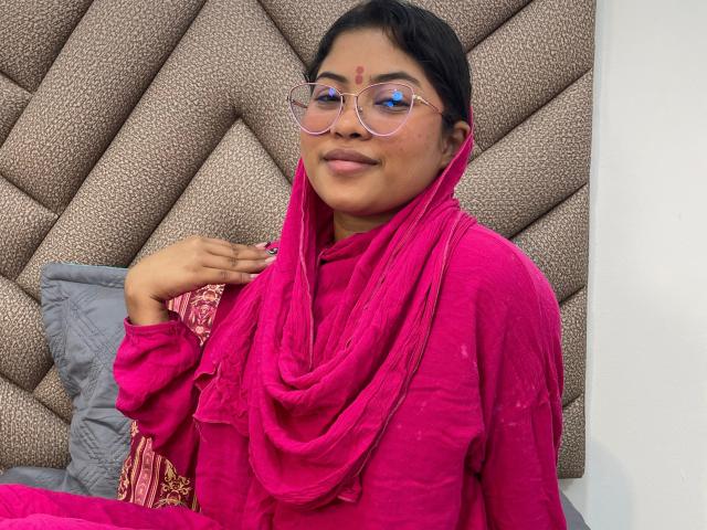 ShailaYojana with dark hair, glasses, and pink traditional outfit, smiling, pink scarf, red dot on forehead, hand near chest, brown patterned headboard, gray pillows, pink and red fabric.