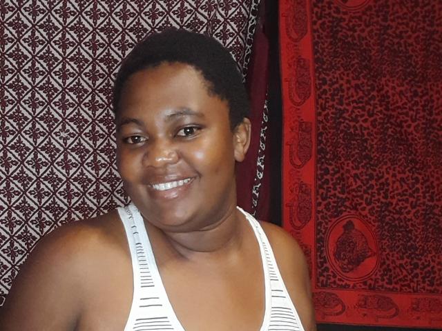 DoraKatty with dark skin, short curly black hair, smiling, wearing a white striped tank top, showing small, cute tits, standing in front of red and black patterned curtains.