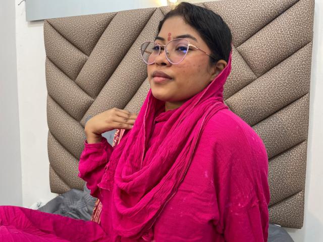 ShailaYojana, wearing a bright pink saree, has glasses on and a red bindi. She's sitting on a bed with a quilted headboard, looking at the camera.