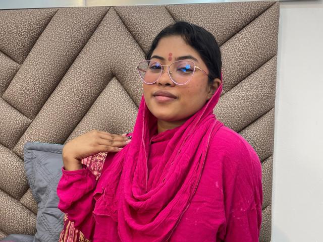 ShailaYojana with medium brown skin, black hair, and pink glasses, wearing a pink dress and matching scarf, has a red dot on her forehead, and a relaxed smile.