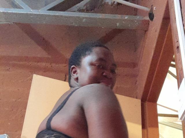 DoraKatty with dark skin, curly hair, and small breasts, wearing a black strap top, looking over her shoulder in a dimly lit room with a metal ceiling.