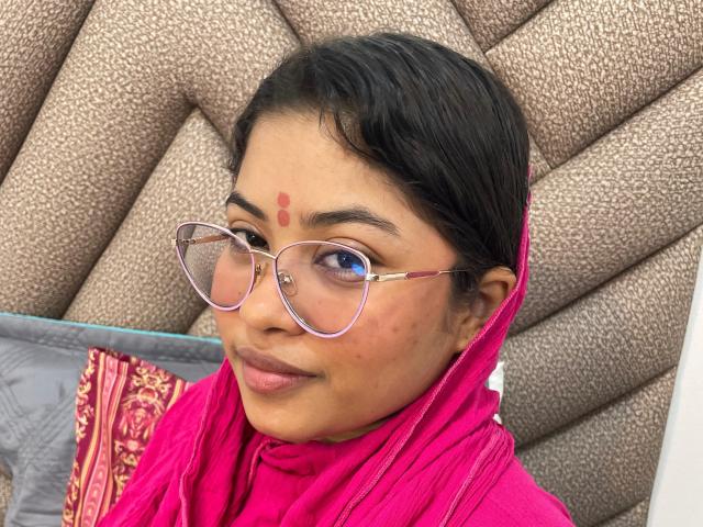 ShailaYojana with dark hair, pink saree, and red bindi, wearing glasses with pink frames, has blue eyes, and a slight smile, showing small acne marks on her right cheek.