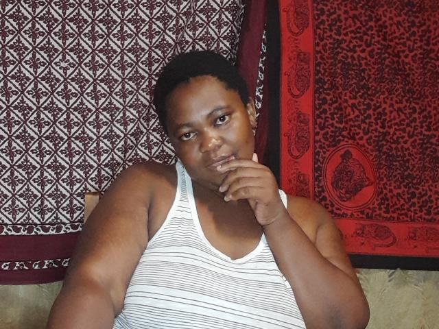 DoraKatty with dark skin, short curly black hair, wearing a white striped tank top, has a chubby build, sits with finger to chin, looking at camera.