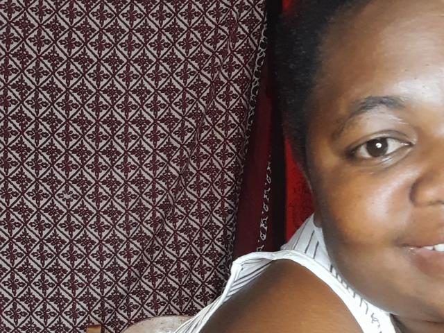 DoraKatty has dark brown skin, short curly black hair, and is smiling slightly, showing her teeth. She's wearing a white sleeveless top, with her right shoulder visible.