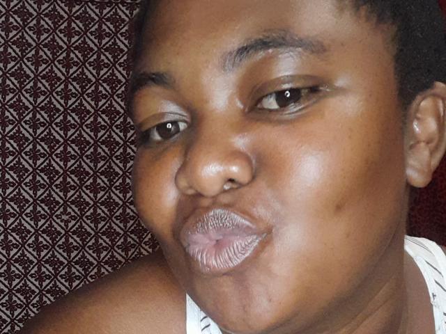 DoraKatty with dark brown skin, puffy lips, and short curly hair, smiling slightly with a nose ring. She's wearing a white top, looking at the camera.