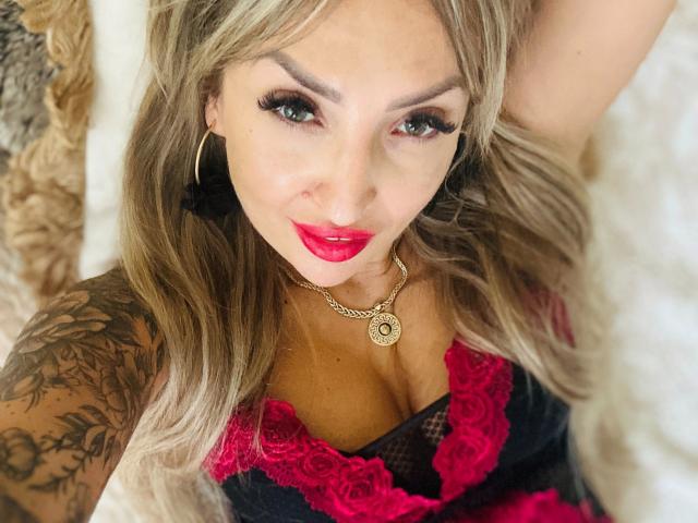LadyKatie-hot with red lipstick, black lace lingerie, tattoos on her arm, and a gold necklace, looking up with green eyes, blonde hair, and black ear cuffs.