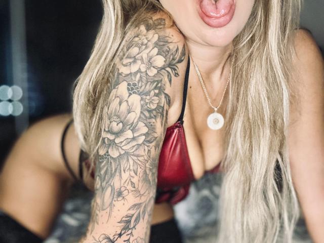 LadyKatie-hot with tongue out, red satin bra, black thong, and tattooed arm, showing large tits and a flower tattoo. She's wearing a necklace with a circular pendant.