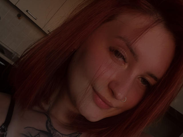 DarkXTemptress with red hair, nose ring, slight smile, looking at camera, showing a tattoo on her chest, dim lighting, visible collarbone.