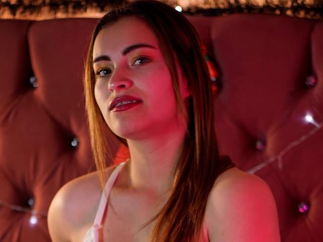 MadissonSutter has straight brown hair, light skin, and is looking to the side with a slight smile. She's wearing a white tank top, showing her bare shoulders and tits.