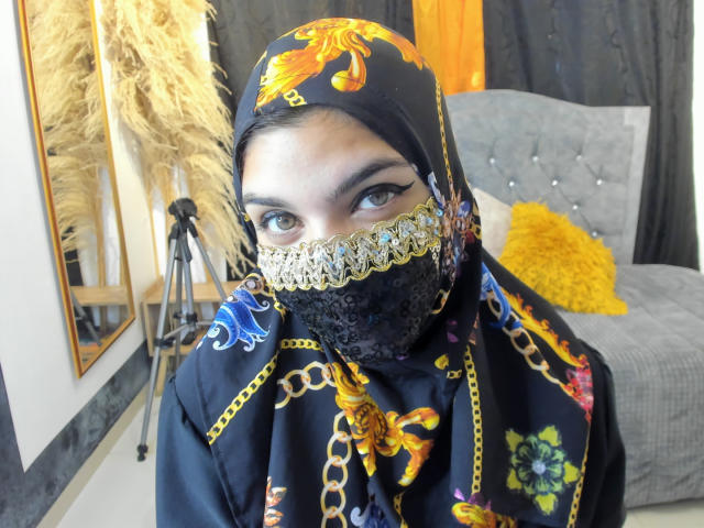 AniisaAli with dark eyes, wearing a black floral hijab, lace mask, and colorful patterned black robe, has a sexy, mysterious look with yellow eyes and thick eyebrows.