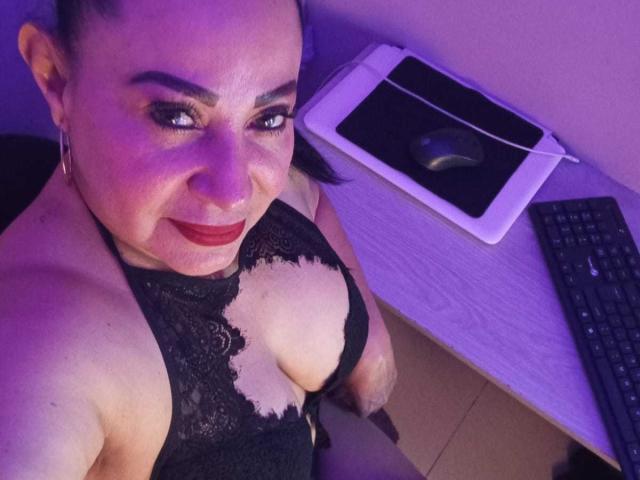 LucianaBlack mit großen Brüsten, trägt ein schwarzes Spitzenoberteil, lächelt in die Kamera, violette Beleuchtung, ein Holztisch im Hintergrund, ein Laptop, eine Maus und eine Tastatur. Ihr roter Lippenstift und ihre Creolen fallen ins Auge..