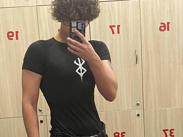 Denjimmie with curly hair, wearing a black t-shirt with a white diamond logo, taking a mirror selfie, holding a phone with a black case, showing a toned, muscular chest.