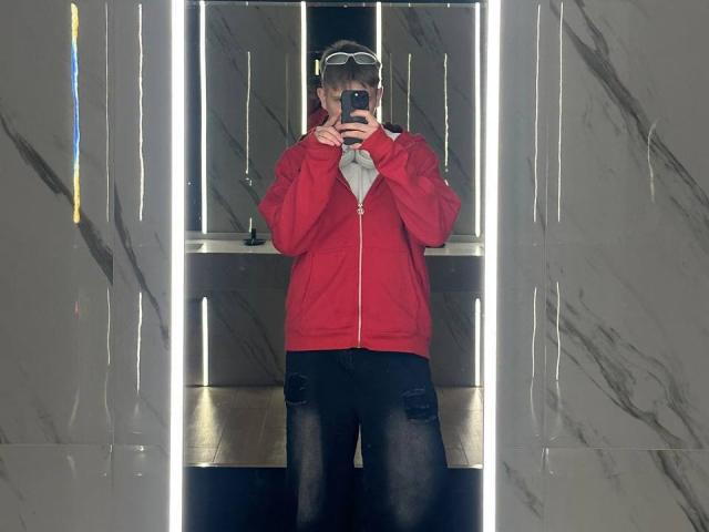 Denjimmie in a red jacket, grey shirt, black pants, and a cap, taking a mirror selfie with a phone, standing in front of vertical LED lights.