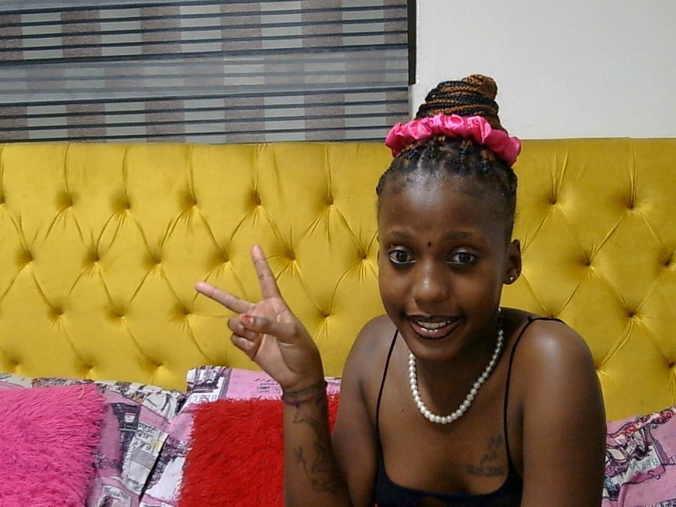 HottieForYou69 with dark skin, braided hair in brown and pink scrunchie, smiling, wearing black top and white necklace, making "peace" sign, tattoos on arms, sitting on yellow tufted bed.