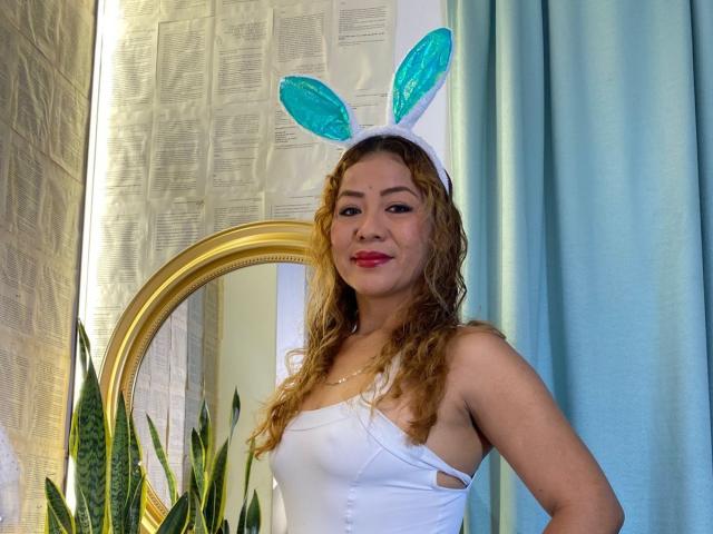 MirandhaX with curly brown hair, wearing a white strapless top and blue-green bunny ears, red lipstick, standing in front of a mirror, blue curtains, and green plants.