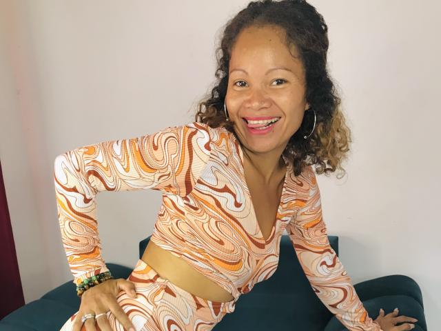 DolloresStone, sorrindo com a boca aberta, vestindo uma blusa cropped e uma saia laranja e branca com padrão de espiral, sentada em um sofá azul-turquesa, cabelos castanhos cacheados, brincos de argola..