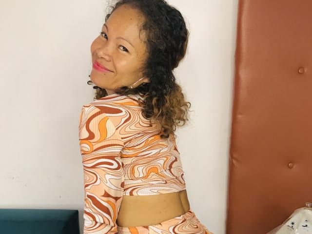 DolloresStone, com cabelos castanhos encaracolados, vestindo uma blusa laranja e branca com padrões, sorrindo para a câmera, mostrando uma cintura fina e usando um fone de ouvido. Os seios não estão visíveis..
