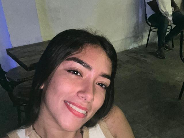 SharonKlart mit dunklem Haar, lächelnd, mit weißen Zähnen, und einem Top ohne Ärmel, ihr kleiner Nasenrücken und ihre vollen Lippen sind sichtbar, sitzend in der Nähe eines Holztisches und eines Stuhls..
