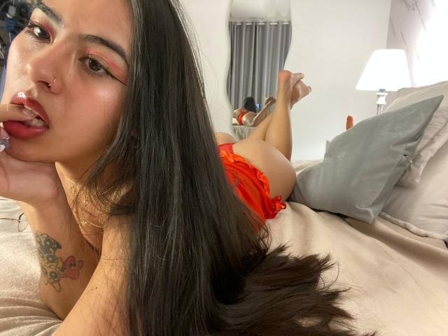 SamanthaRosses with long black hair, red lipstick, and nose ring, lying on a bed in red lingerie, sucking her finger, showing a tattoo on her arm, and a butt.