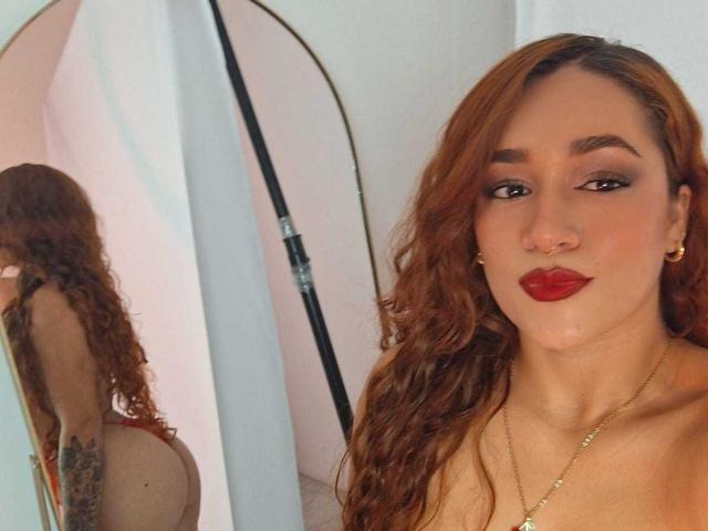 Valery Hilton mit rotem Lippenstift und welligem, braunem Haar, die in einen Spiegel schaut, der ihren nackten Rücken und ein Tattoo zeigt, trägt eine goldene Halskette und Ohrringe und hat leicht geschwollene Lippen..