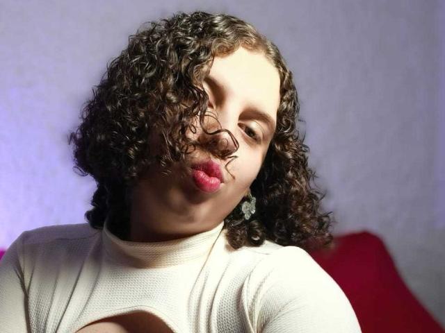 CatleyaNoire, met krullend bruin haar, roze lippenstift, een witte gebreide top en diamanten oorbellen, zet haar volle, ronde, lichte lippen op een frons, waarbij een subtiele decolleté zichtbaar is..