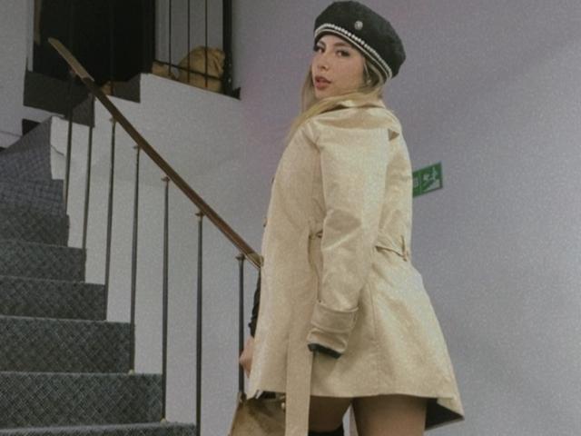 AmaraRicci, wearing a beige trench coat and gray beanie, stands on carpeted stairs, showing her ass, with a brown handbag and a confident, sexy look.