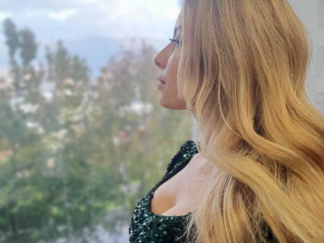 AmaraRicci, avec de longs cheveux blonds ondulés, vue de profil, portant un haut vert pailleté, regardant par une fenêtre, laissant légèrement entrevoir ses seins à cause du décolleté plongeant du haut..