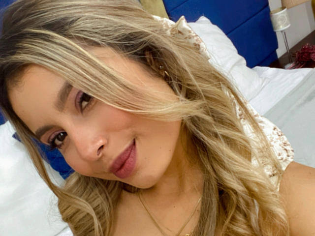 AmaraRicci has blonde, wavy hair, light brown eyes, and full pink lips, wearing a lace top, with a subtle smile and a gold necklace, in a bedroom.