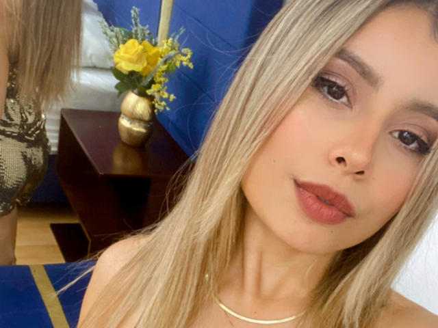 AmaraRicci has blonde hair, full lips with pink lipstick, and a small nose. She's wearing a gold necklace, and her brown eyes are slightly open in a relaxed look.