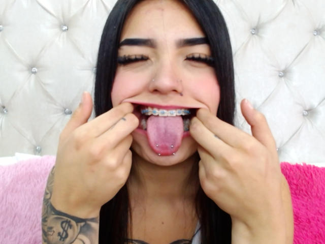 MiaXSmithy with black hair, nose ring, and tongue piercing, sticking out her tongue, showing metal braces, with hands on her cheeks, tattoo on right arm, smiling.