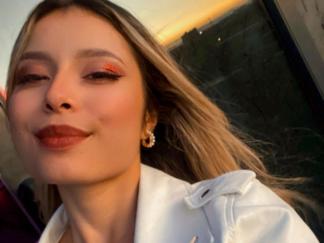Amara Ricci, com cabelo loiro, vestindo um casaco branco, com sombra laranja e batom marrom, sorrindo levemente, brincos de ouro, luz do sol no rosto..